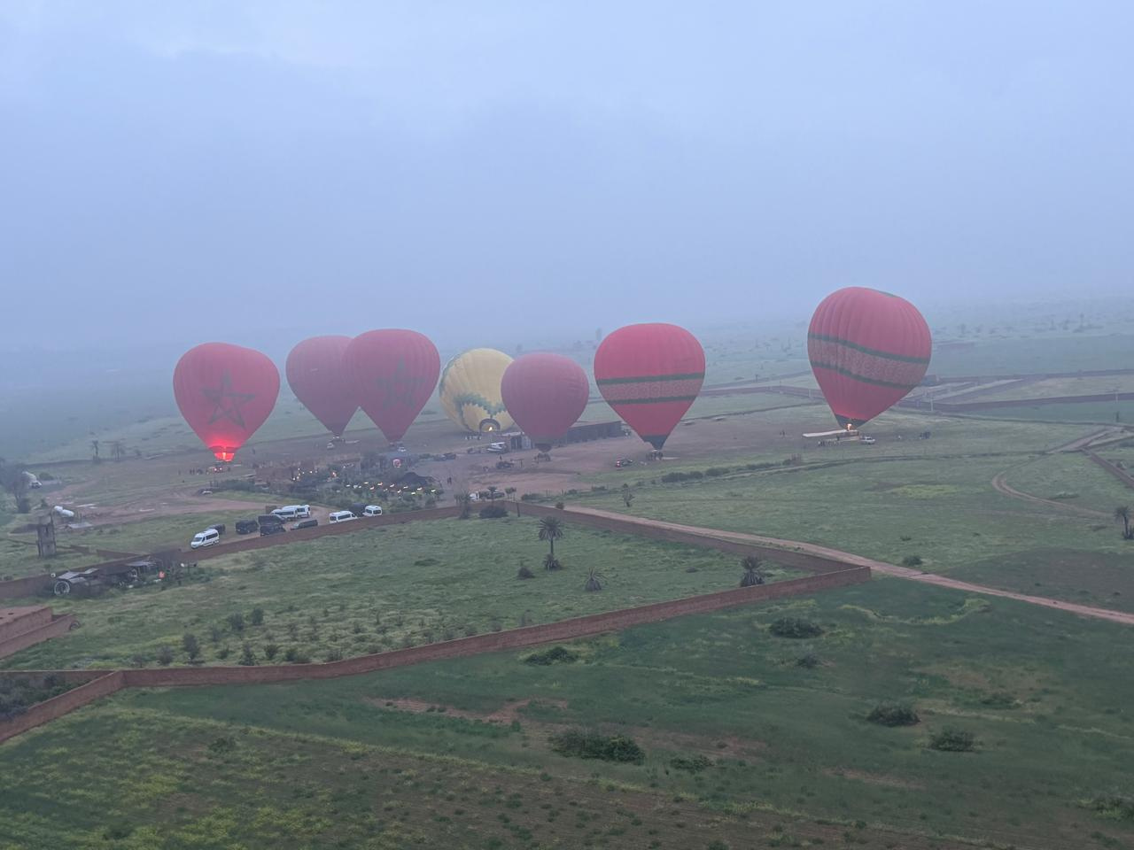Several colorful hot air balloons flying above green fields and rural tracks near Marrakech