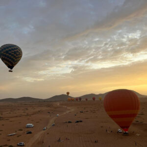 Several hot air balloons over a launch field with vehicles beneath a dramatic sunrise sky near Marrakech