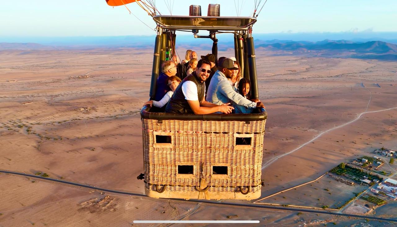 Passengers leaning from a wicker hot air balloon basket above fields and roads near Marrakech