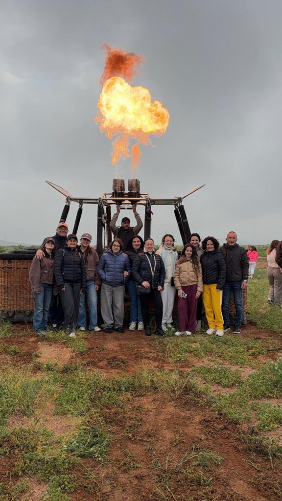 Travelers gathered beside a hot air balloon basket while a large burner flame fires upward near Marrakech