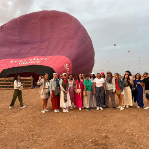Travel group posing in front of a large pink hot air balloon envelope on a launch field near Marrakech