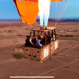 Passengers in a hot air balloon basket with bright burner flames firing above open plains near Marrakech