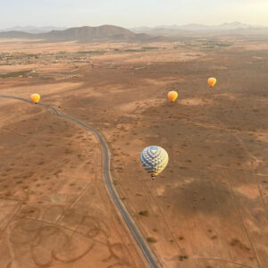 Several colorful hot air balloons flying above a road across broad ochre plains near Marrakech