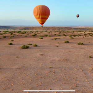 Several hot air balloons flying across open scrubland with one orange balloon in front near Marrakech