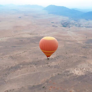 Orange hot air balloon flying over wide dry plains with distant mountains on the horizon near Marrakech