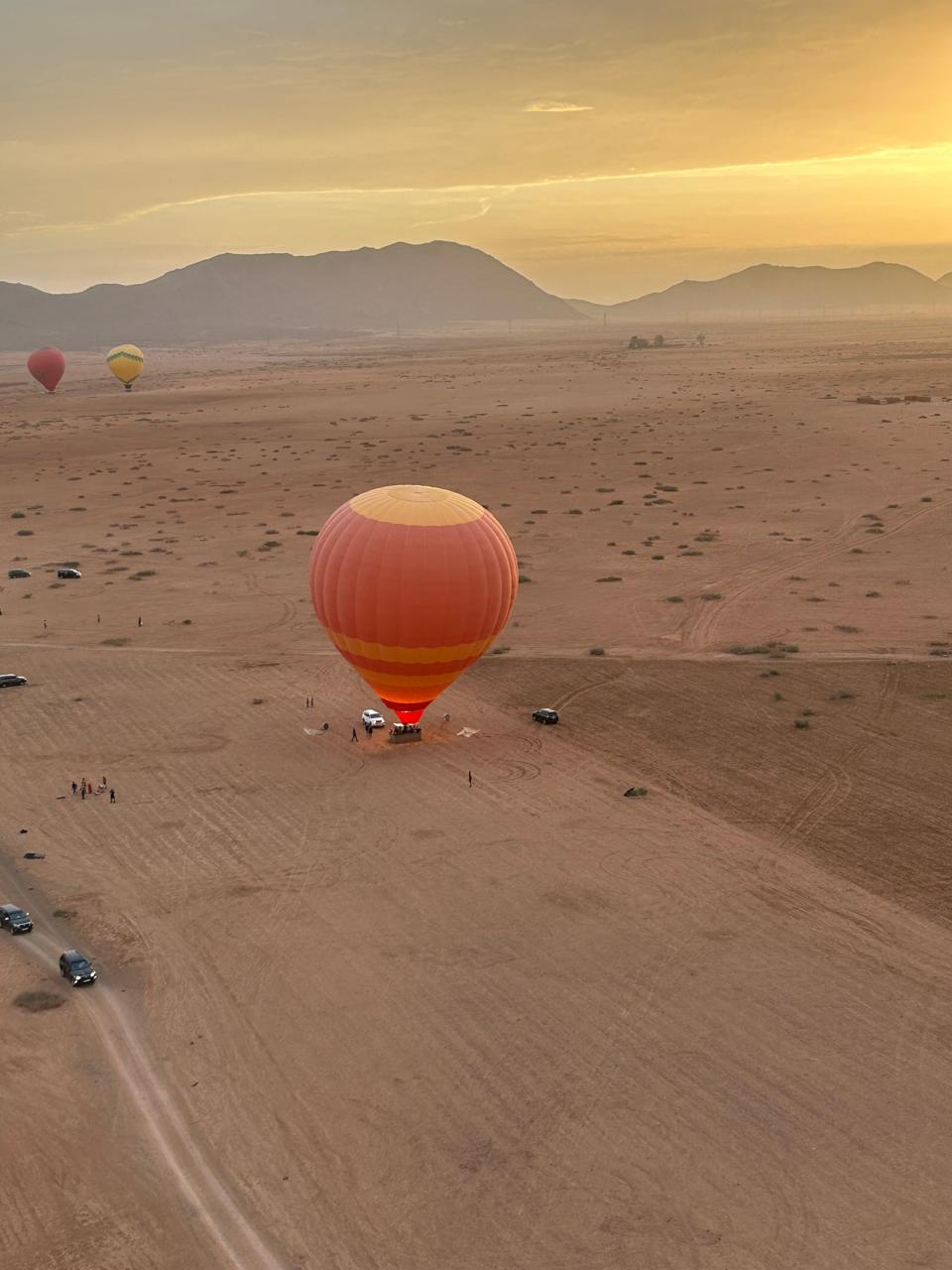 Orange hot air balloon flying over open terrain toward a glowing sunrise and distant mountains near Marrakech