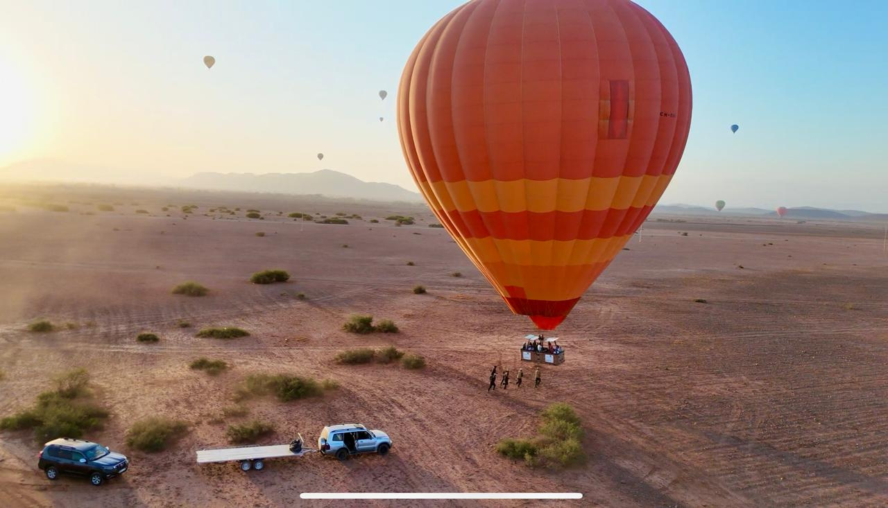 Orange hot air balloon flying low above open ground with support vehicles waiting near Marrakech