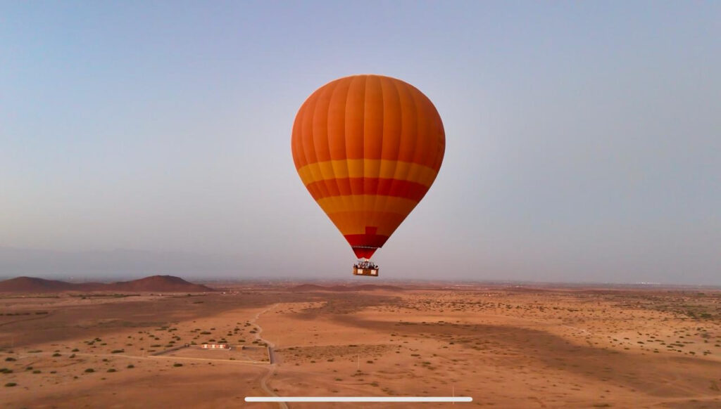 Orange hot air balloon flying low over tan countryside with tracks and sparse shrubs near Marrakech