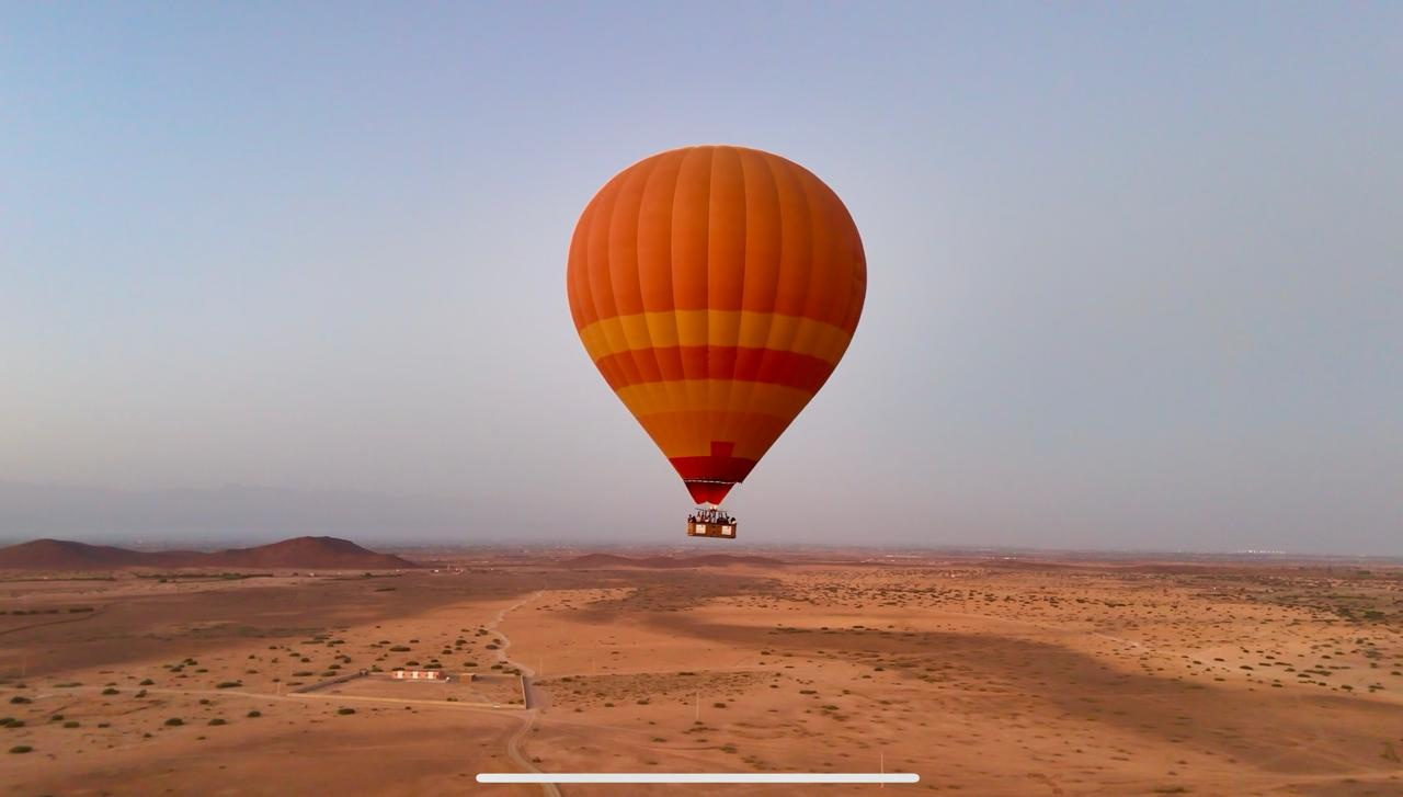 Orange hot air balloon flying low over tan countryside with tracks and sparse shrubs near Marrakech