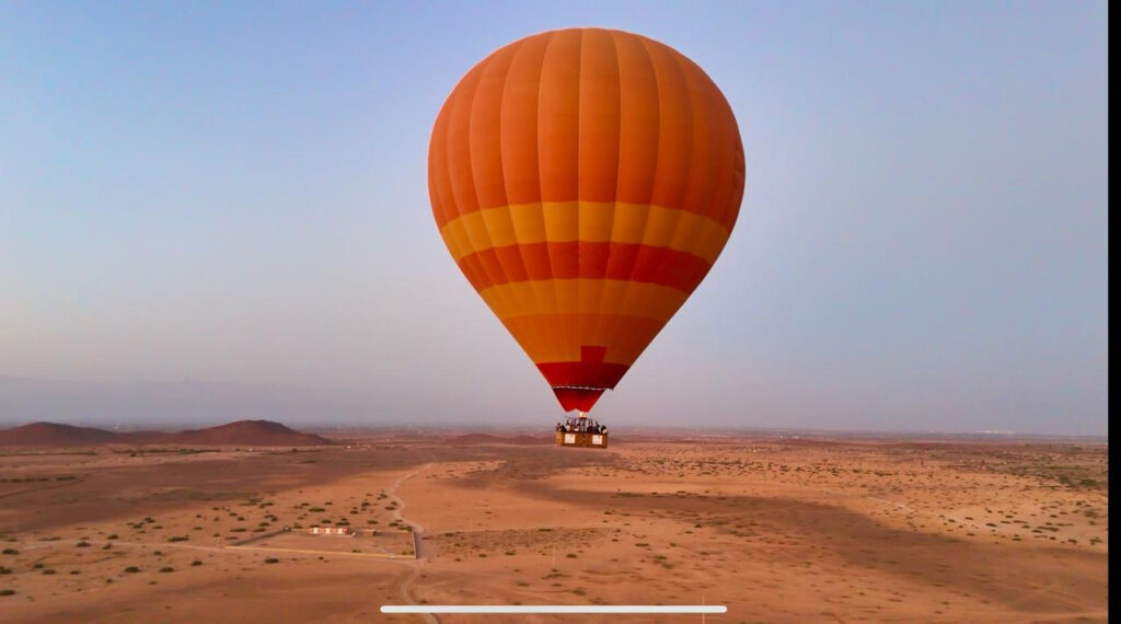 Bright orange hot air balloon floating above sandy open terrain during a morning flight near Marrakech