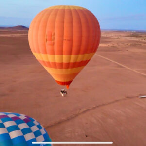 Large orange hot air balloon flying over an arid plain with tracks and sparse vegetation near Marrakech