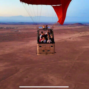 Hot air balloon basket with passengers floating above a broad dry landscape and roads near Marrakech