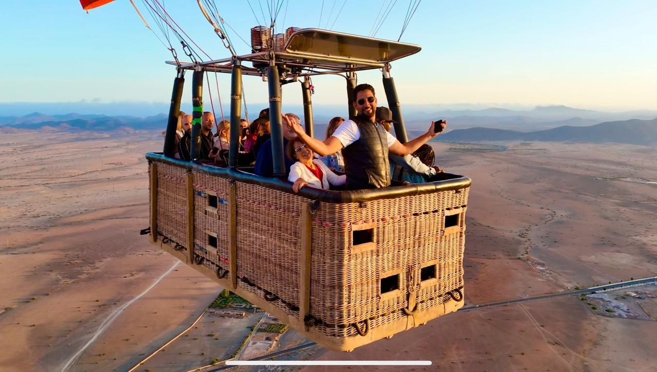 Smiling passengers waving from a wicker hot air balloon basket over the countryside near Marrakech