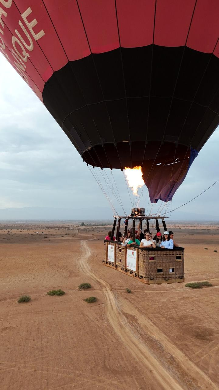 Red and black hot air balloon carrying passengers low above a dirt track near Marrakech