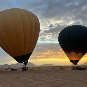 Two large hot air balloons near the ground beneath a dramatic cloud-filled sunrise sky near Marrakech